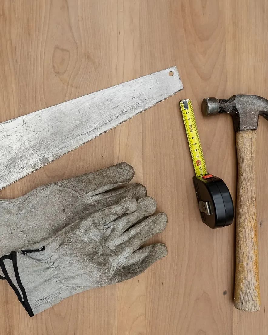 Symbolbild: Säge, Arbeitshandschuhe, Maßband und Hammer auf einer Holzplatte